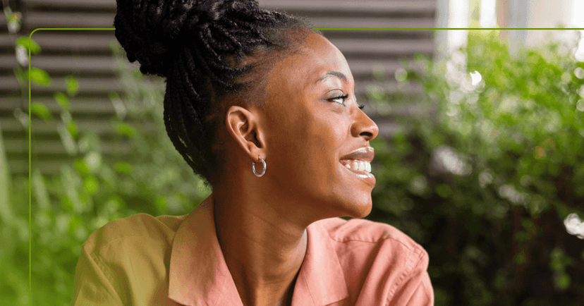 A woman looks off to the left with a satisfied smile on her face. There are plants in the background.