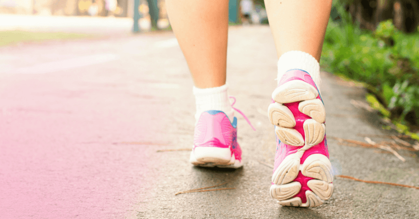 Close up photo of two legs wearing athletic shoes while walking on a sidewalk. The shoes are pink and white.