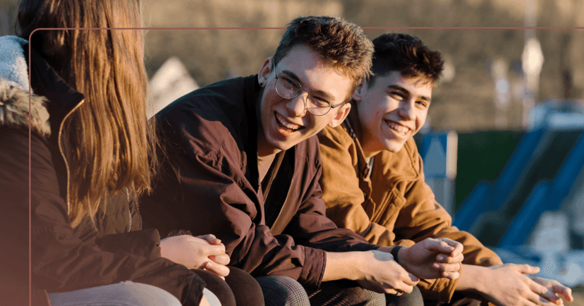 Two young men sit outside next to a young woman on the left facing away from the camera. They are laughing and the facial expression of the young woman is not visible. They are all wearing coats and the background is a parking lot.