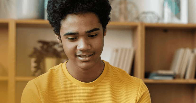 A male teenager is looking straight down off camera. There is a bookshelf visible in the background. He is wearing wearing a yellow shirt.