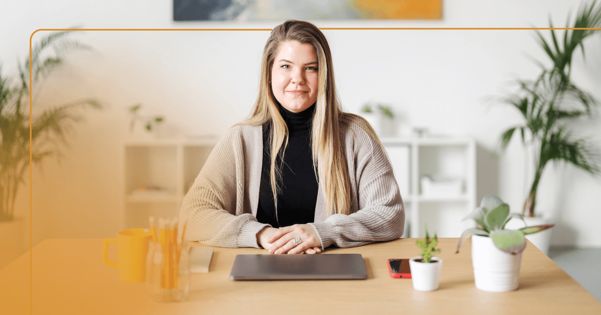 Woman sits at a tan wooden table with a closed laptop in front of her staring straight into the camera. She has a smirk and her hands crossed in front of her on the table. She is wearing a black turtleneck and tan sweater. The background is green plants against a white wall.