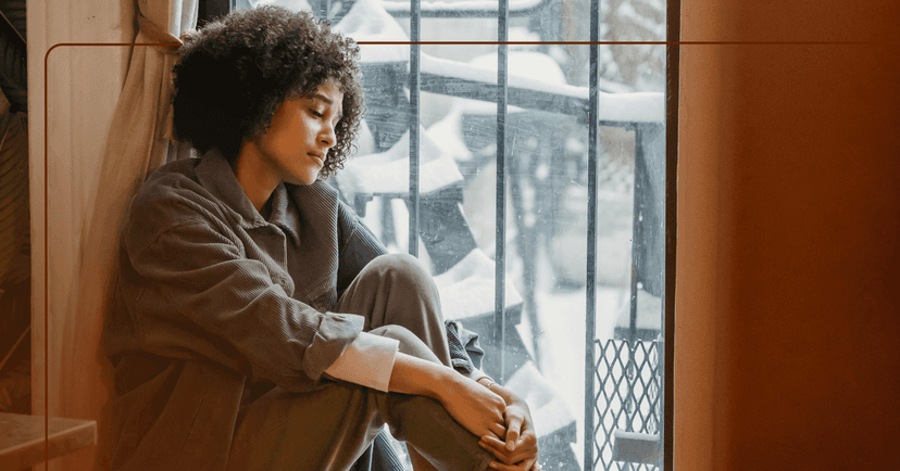 Woman sits on the floor next to a window. She is holding her bent knees and looking down. Her facial expression is sad. She is wearing a gray long sleeve shirt and gray pants. There are black metal bars on the window.