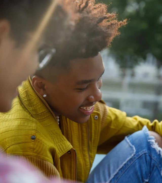 Smiling person with curly hair in a yellow jacket and ripped jeans, sitting outdoors. Another person is partially visible in the foreground.