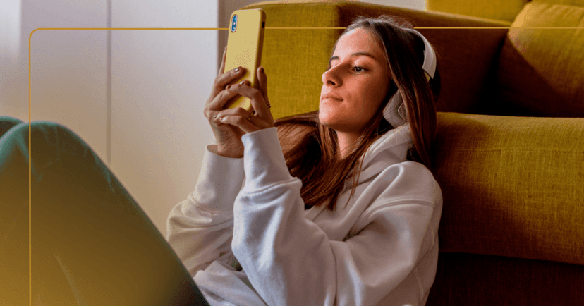 Young girl sits on the floor leaning against a yellow couch. She is looking at the yellow phone she holds in front of her in both hands. She is wearing white over the ear headphones and a white hoodie. There is a yellow border around the image.