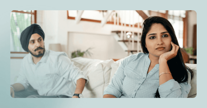 Couple sits on a couch downstairs in their home. The man is on the left looking at the woman. His facial expression is angry. The woman is on the right leaning on her left hand staring off. She is wearing a blue button-up blouse and is sharper in focus.