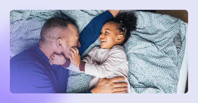 Dad and child lay in a bed. They are looking at each other and smiling, maybe playing. The background is a blue patterned duvet cover. The girl has her hand on her dad's face.