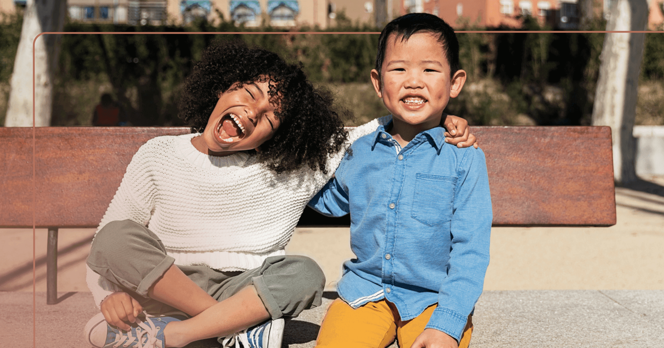 Two boys sit arms in arms on an outdoor concrete bench. One has a small open smile and the other appears to be yelling happily.