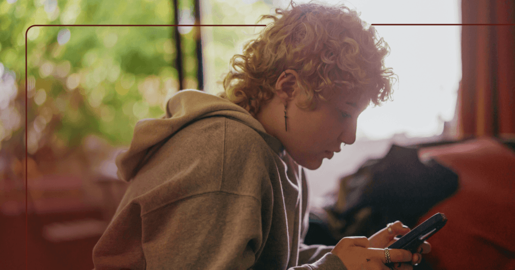 Young woman sits in the center of the screen. They are looking down at their cellphone and typing with both hands. They are wearing a gray hoodie with earrings. The background is a bedroom with items on the floor.