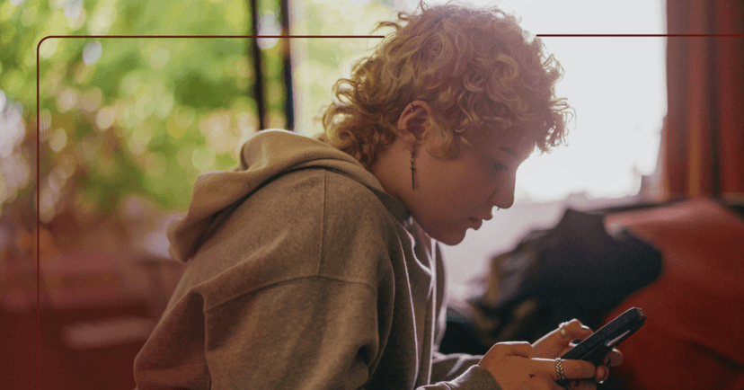 Young woman sits in the center of the screen. They are looking down at their cellphone and typing with both hands. They are wearing a gray hoodie with earrings. The background is a bedroom with items on the floor.
