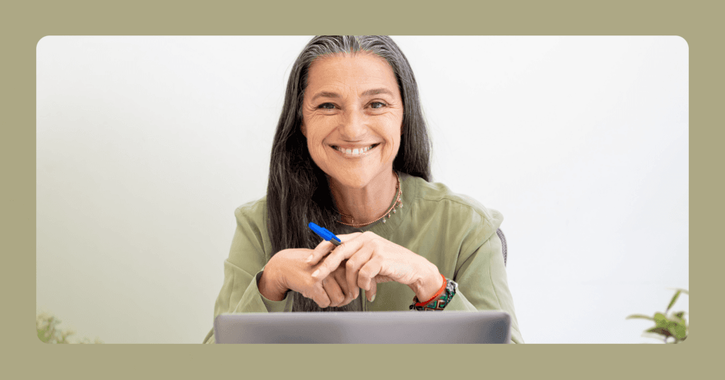 Older woman sits staring straight into the camera and smiling with her teeth. There is an open laptop in front of her and a blue pen in her left hand. She is wearing a green long sleeve shirt and a necklace. There is a green border around the image that matches her shirt. The background is a white wall.