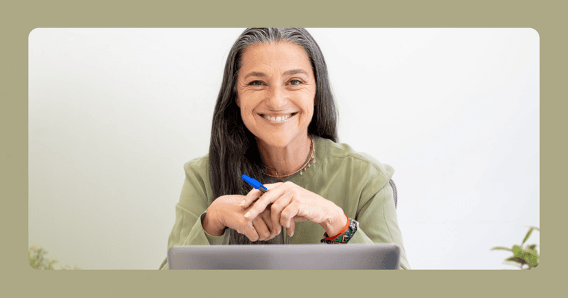 Older woman sits staring straight into the camera and smiling with her teeth. There is an open laptop in front of her and a blue pen in her left hand. She is wearing a green long sleeve shirt and a necklace. There is a green border around the image that matches her shirt. The background is a white wall.