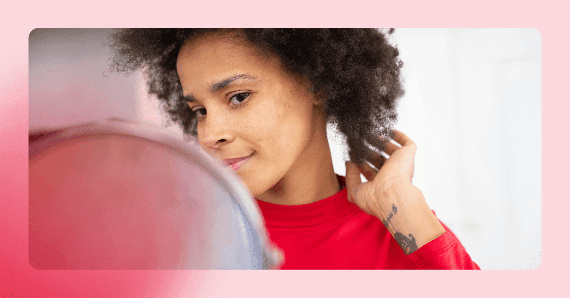 Woman with dark, curly hair is looking into a mirror with a slight smirk. She is wearing a red long sleeve shirt.
