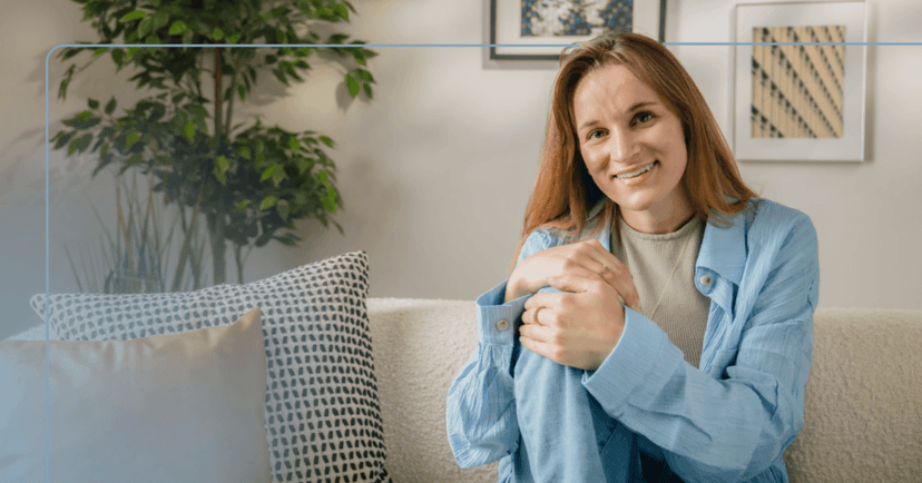 Woman sits on a white couch staring straight into the camera. She is wearing a gray shirt underneath a light blue button up. She is smiling with her teeth and holding her left knee. The background is a green plant and two picture frames against a white wall.