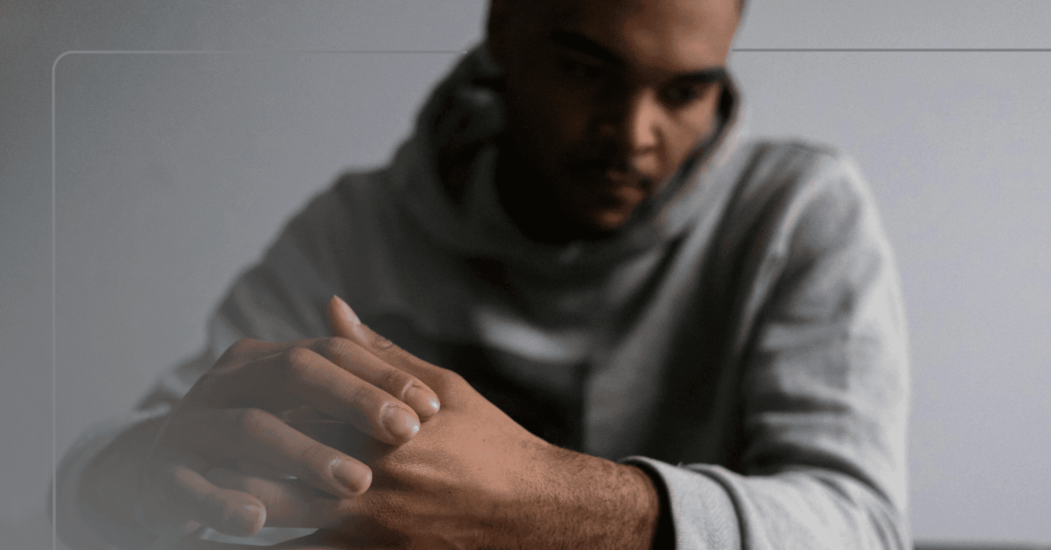 A man in a grey hooded sweatshirt sits with his hands held together. He looks off camera to the left. There is a gray wall in the background.