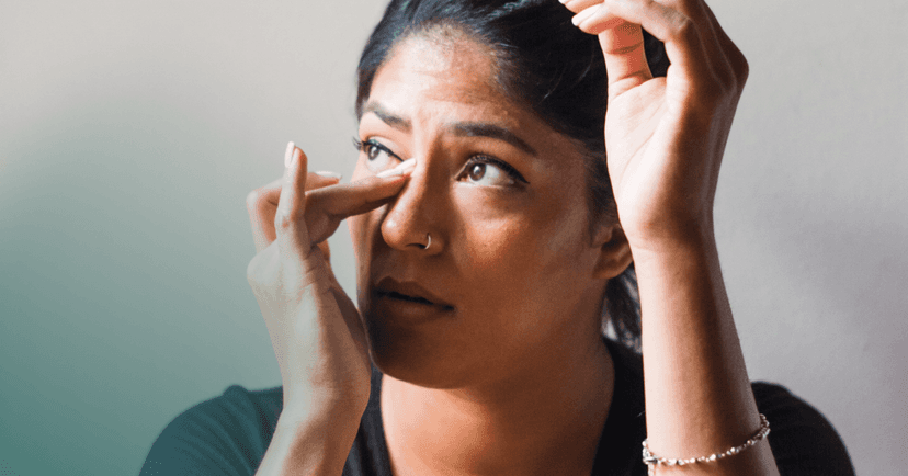 Woman is looking away with a sad expression. Her right hand is wiping away a tear. She is wearing a black shirt and the background is a plain wall.