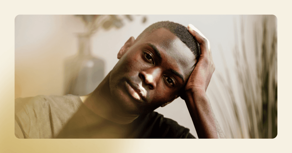 Young man stares straight into the camera with his head turned to the left against his left hand. His facial expression is serious and distraught. He is wearing a black T-shirt. The background is two plants.