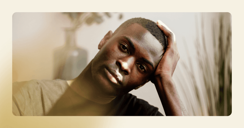 Young man stares straight into the camera with his head turned to the left against his left hand. His facial expression is serious and distraught. He is wearing a black T-shirt. The background is two plants.