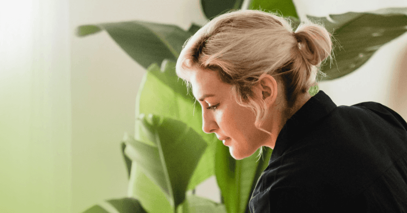 A woman with blonde hair in a bun stares off the left side of the frame. She is wearing a black shirt and standing in front of a large green house plant.