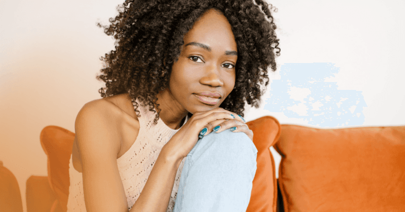 Woman sits on an orange couch staring straight into the camera. She leans on both her hands over an extended knee. She is wearing a crochet tank top, light blue jeans, and teal nail polish. The background is a white wall.