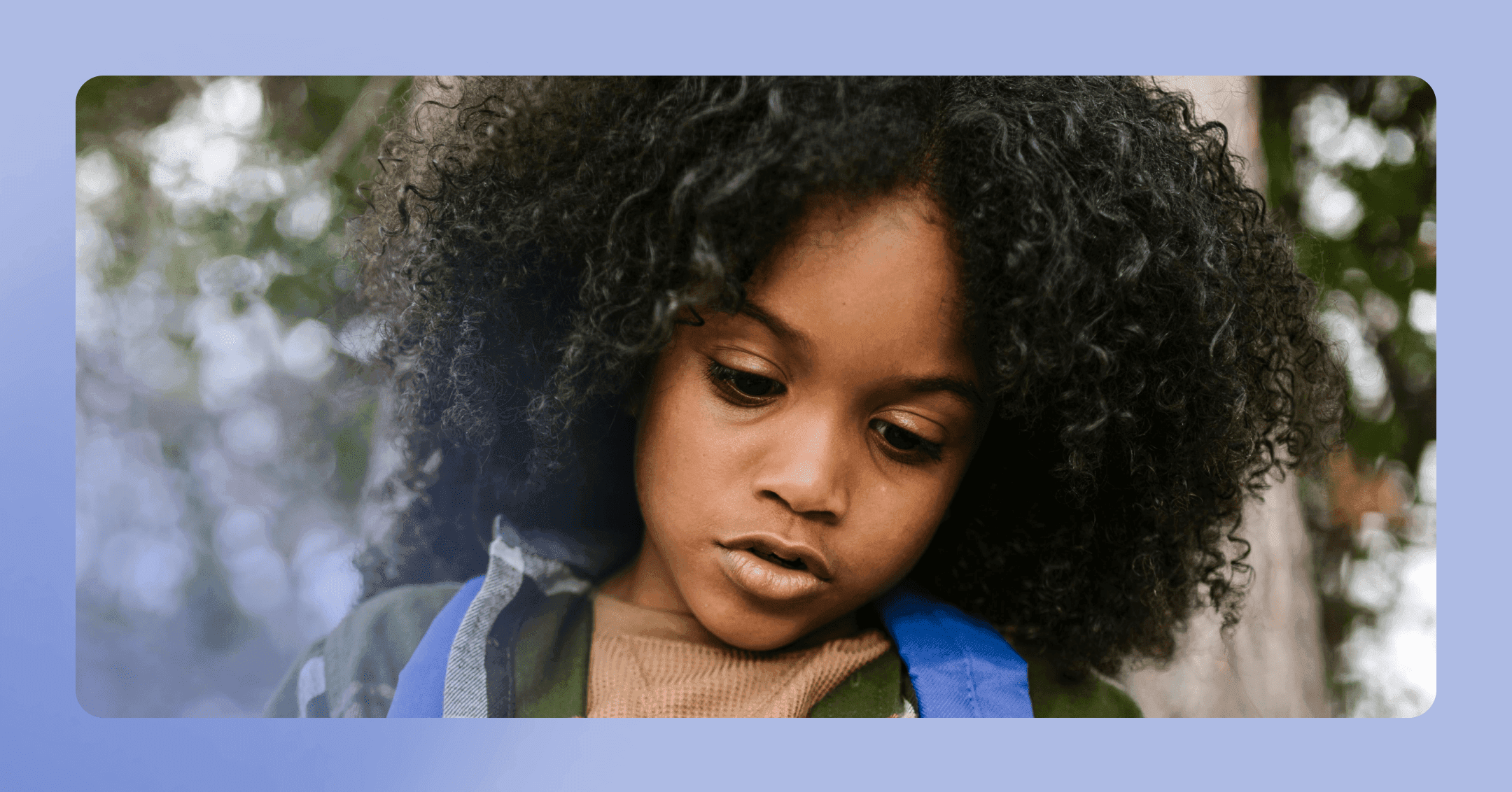 A boy with long, black curly hair looks down with a neutral look on his face. His wearing a blue backpack.