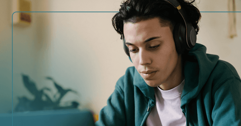 A teenager in a green hoodie with dark hair and headphones on looks down at a laptop with a neutral face. A plant is out of focus in the background.