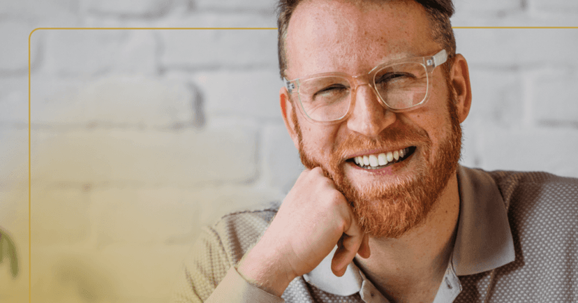 Man sits looking straight at the camera smiling with all his teeth. He is wearing glasses and a gray long sleeve shirt. He is leaning on his right hand in a fist under his chin. The background is a white wall.