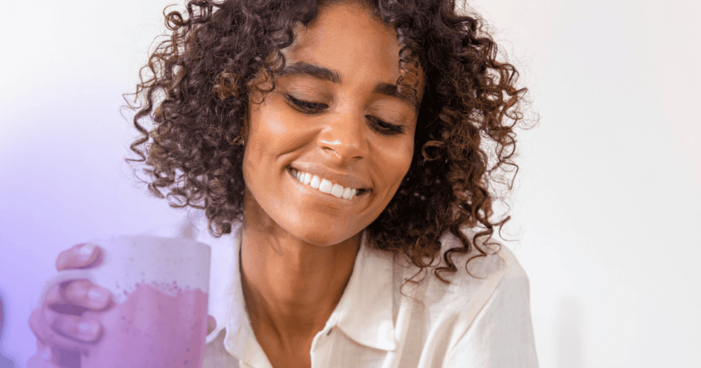 Woman with a cup of coffee or tea in her hand looks down off camera with a smile and closed eyes.