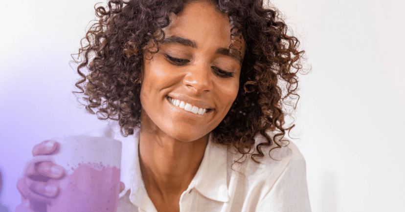 Woman with a cup of coffee or tea in her hand looks down off camera with a smile and closed eyes.
