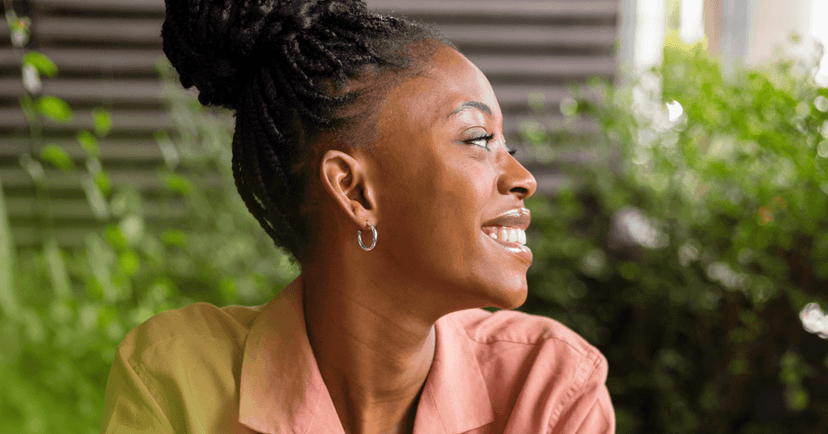 Woman is sitting looking at something offscreen to the left. She is smiling with her teeth. She is wearing a pink button up shirt and small silver hoop earrings. The background is green bushes against a building outside.
