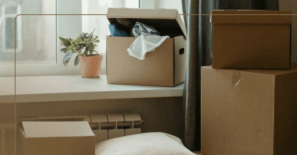 Image shows two brown cardboard moving boxes closed with tape on the right. On the left are two smaller open brown boxes with T-shirt spilling out of one box. The boxes are by a bedroom window. There is a small plant beside one of the open boxes.