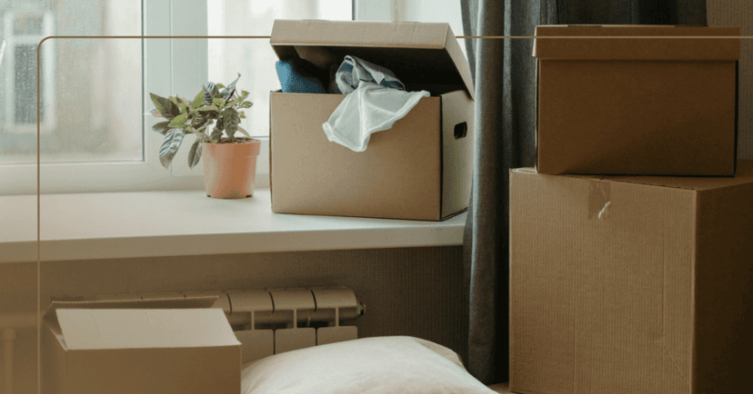 Image shows two brown cardboard moving boxes closed with tape on the right. On the left are two smaller open brown boxes with T-shirt spilling out of one box. The boxes are by a bedroom window. There is a small plant beside one of the open boxes.