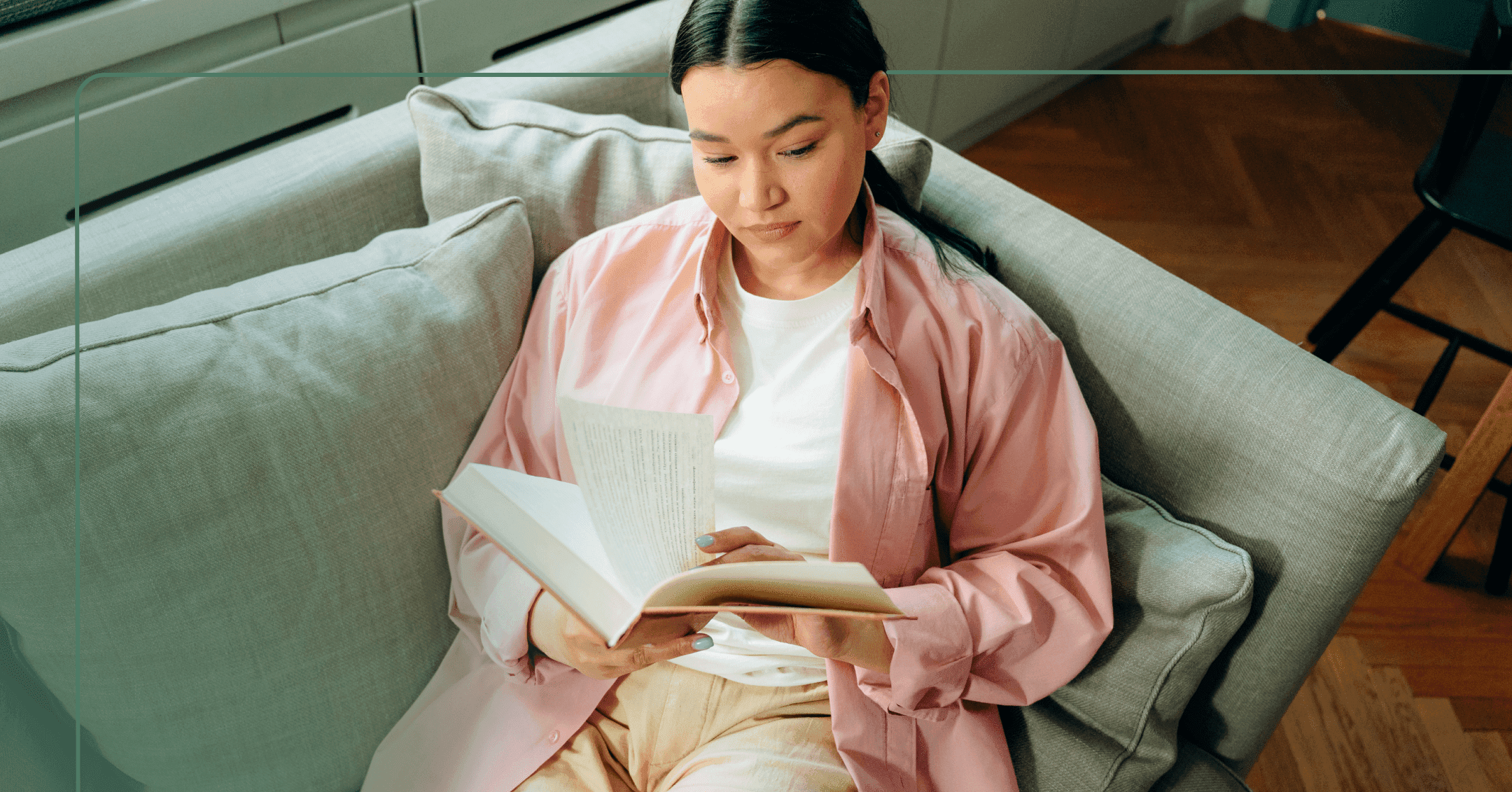 Woman sits on a couch flipping through pages of a hardcover book. She is looking down at the page she is reading. She is wearing a pink button up shirt over a white T-shirt with tan pants. The background is the couch on a brown hardwood floor.
