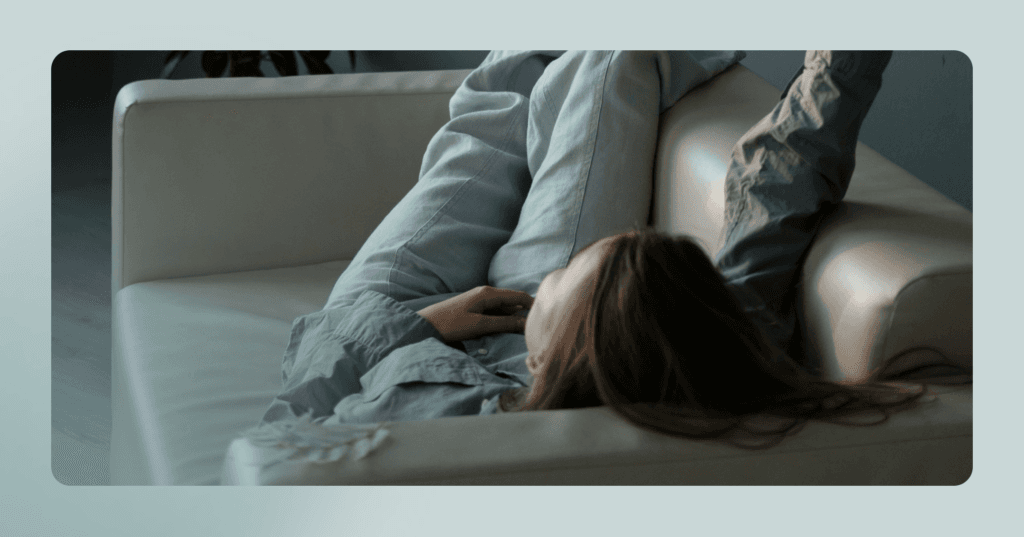 Woman lies down on a white leather couch. Her facial expression is not visible as she faces away from the camera. She is wearing a blue button-up shirt and jeans. Her right arm is raised in the air.