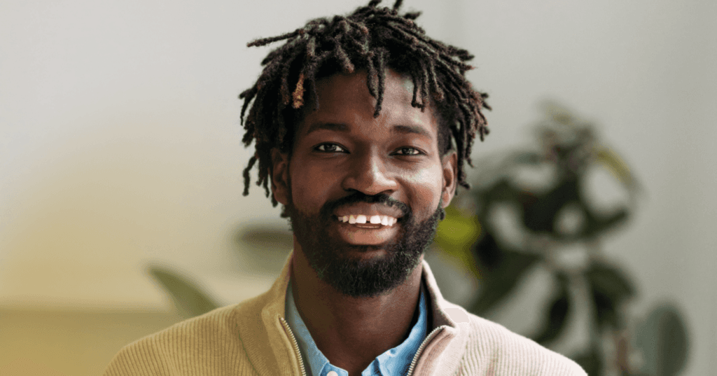 Man stands staring straight into the camera. His facial expression is happy and he is smiling with all this teeth. He is wearing a tan jacket over a blue button-up shirt. The background is a blurry green plant against a white wall.