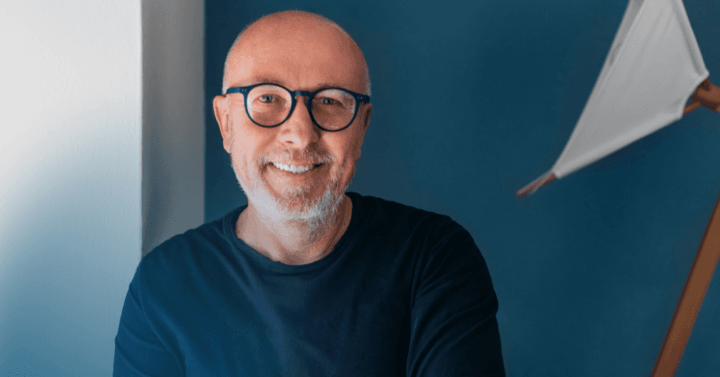 Older man sits staring straight into the camera. He is smiling while wearing a dark blue long sleeve shirt and matching blue glasses. The background is a blue wall that is a shade lighter.