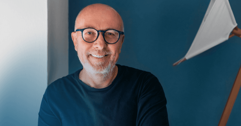 Older man sits staring straight into the camera. He is smiling while wearing a dark blue long sleeve shirt and matching blue glasses. The background is a blue wall that is a shade lighter.