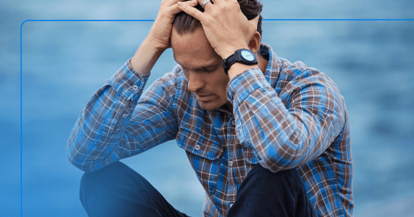 Man in blue and red plaid button up has his head in is hands and an upset look on his face. The ocean is blurry and visible in the background. The image is framed in light blue.