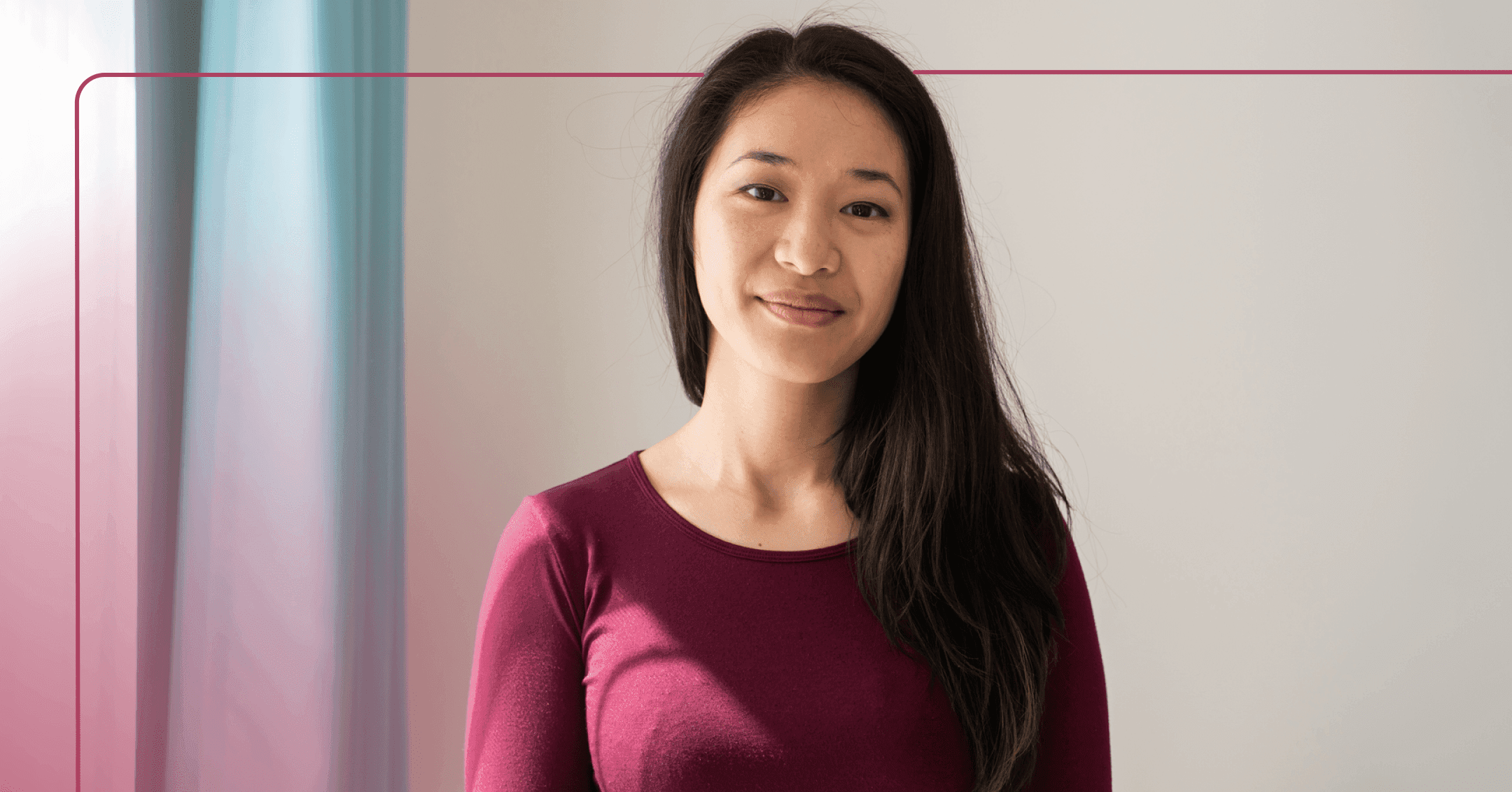 A woman looks directly at camera with a slight smile. She is wearing a maroon long sleeve shirt and is standing if front of a white wall.