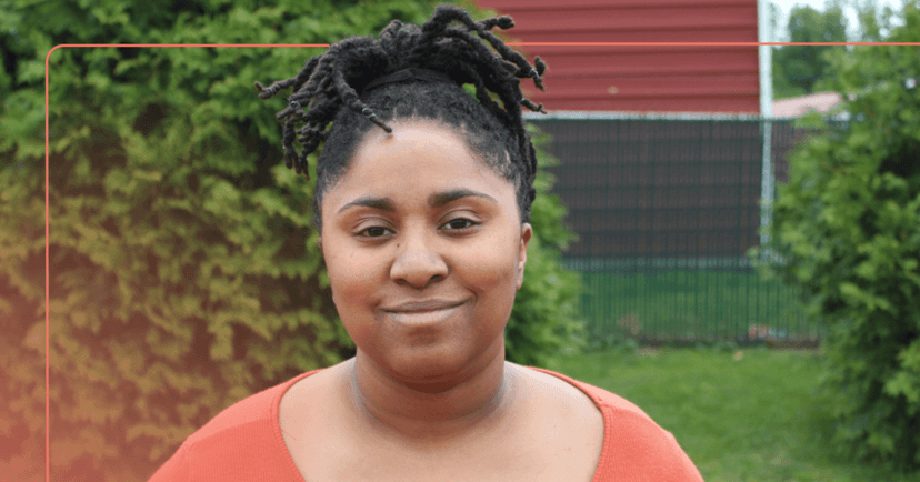 Woman stands outside staring straight at the camera. She is smiling. She is wearing an orange long sleeve shirt. The background is a backyard with trees.