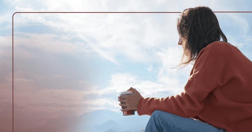 A woman in an orange sweater sits with her hands on her knees and looks out into a visible partly cloudy sky. She is holding a takeout coffee cup.