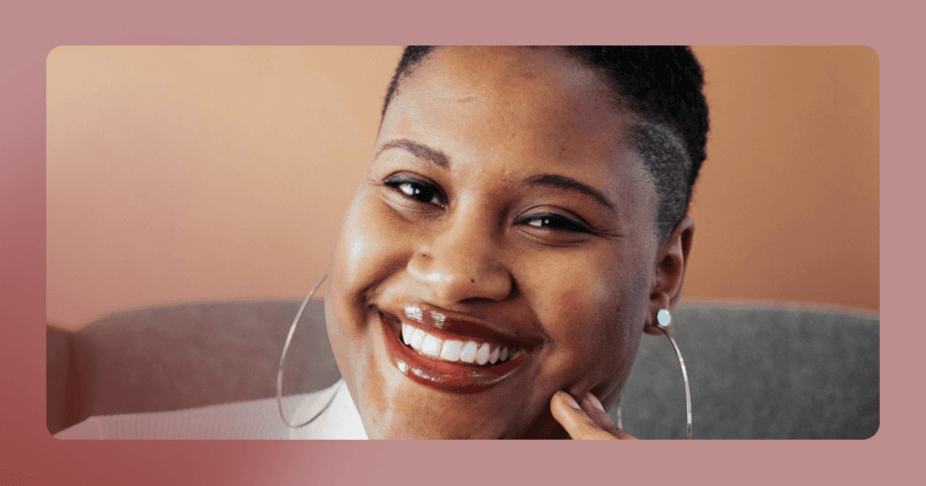 Woman sits on a gray couch staring straight into the camera. She is smiling with all her teeth. She is wearing a white long sleeve shirt and silver hoop earrings. The background is an orange wall behind the couch.