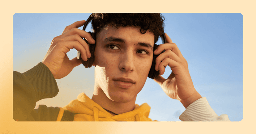 Young man is staring to the right while holding black over the ear headphones with both hands. His facial expression is concerned. He is standing outside wearing a yellow, green, and white hoodie. The background is a clear blue sky.