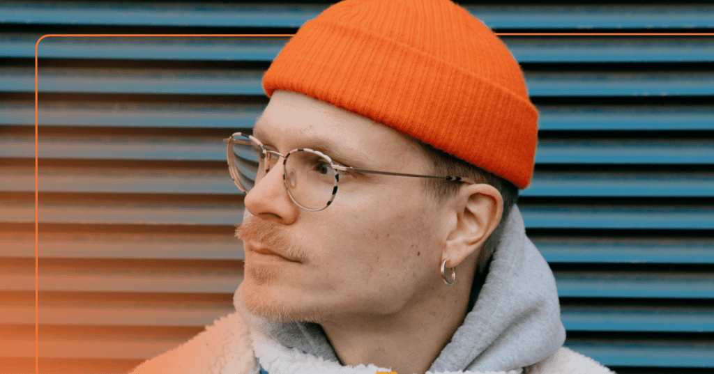 Man with orange beanie and glasses looks off camera to the right. He is standing against a blue metal textured wall. There is an orange frame around the image.