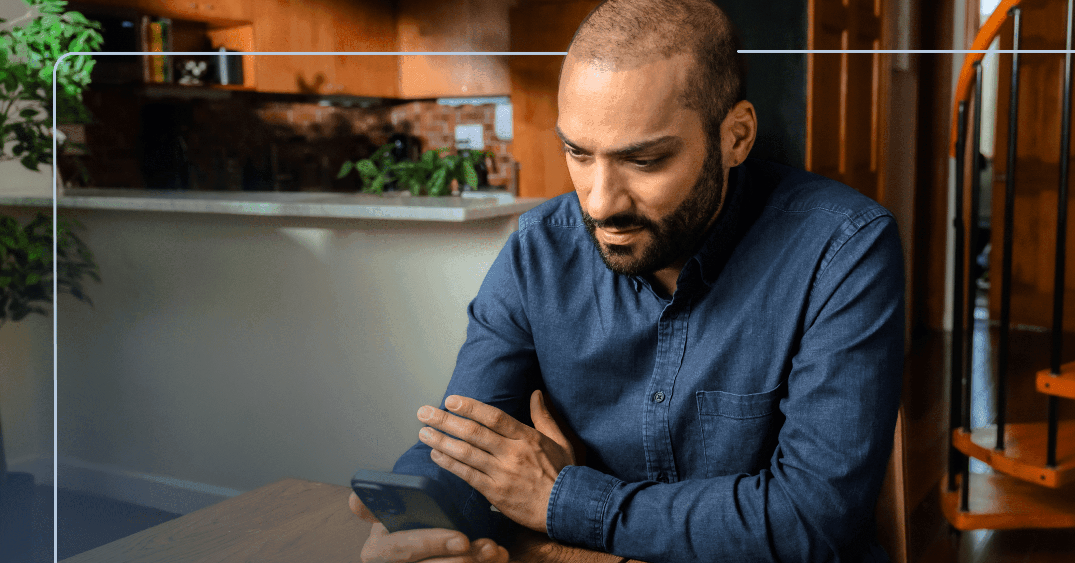 Man sits at a wooden table in his home staring down at his phone in his right hand. His facial expression is neutral. He is wearing a blue denim button up shirt. The background is a kitchen to the left and a staircase on the right.