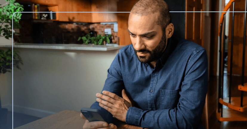 Man sits at a wooden table in his home staring down at his phone in his right hand. His facial expression is neutral. He is wearing a blue denim button up shirt. The background is a kitchen to the left and a staircase on the right.