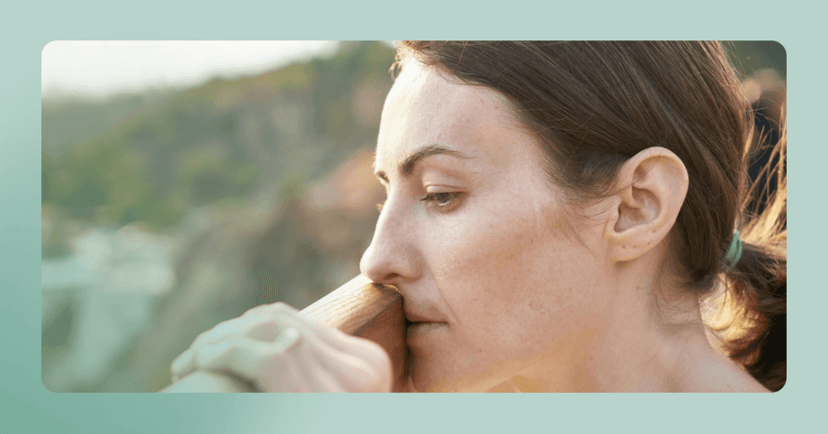 Woman stands outside looking down and leaning against a wooden fence. She is looking offscreen and gripping the fence with her left hand. Only her head and neck are visible. The background is hills.