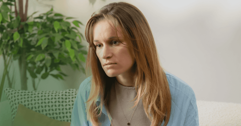 Woman sits on a white couch staring vacantly to the right. She is wearing a gray shirt underneath a light blue button up. Her facial expression is dazed and exhausted. The background is a green plant against a white wall.
