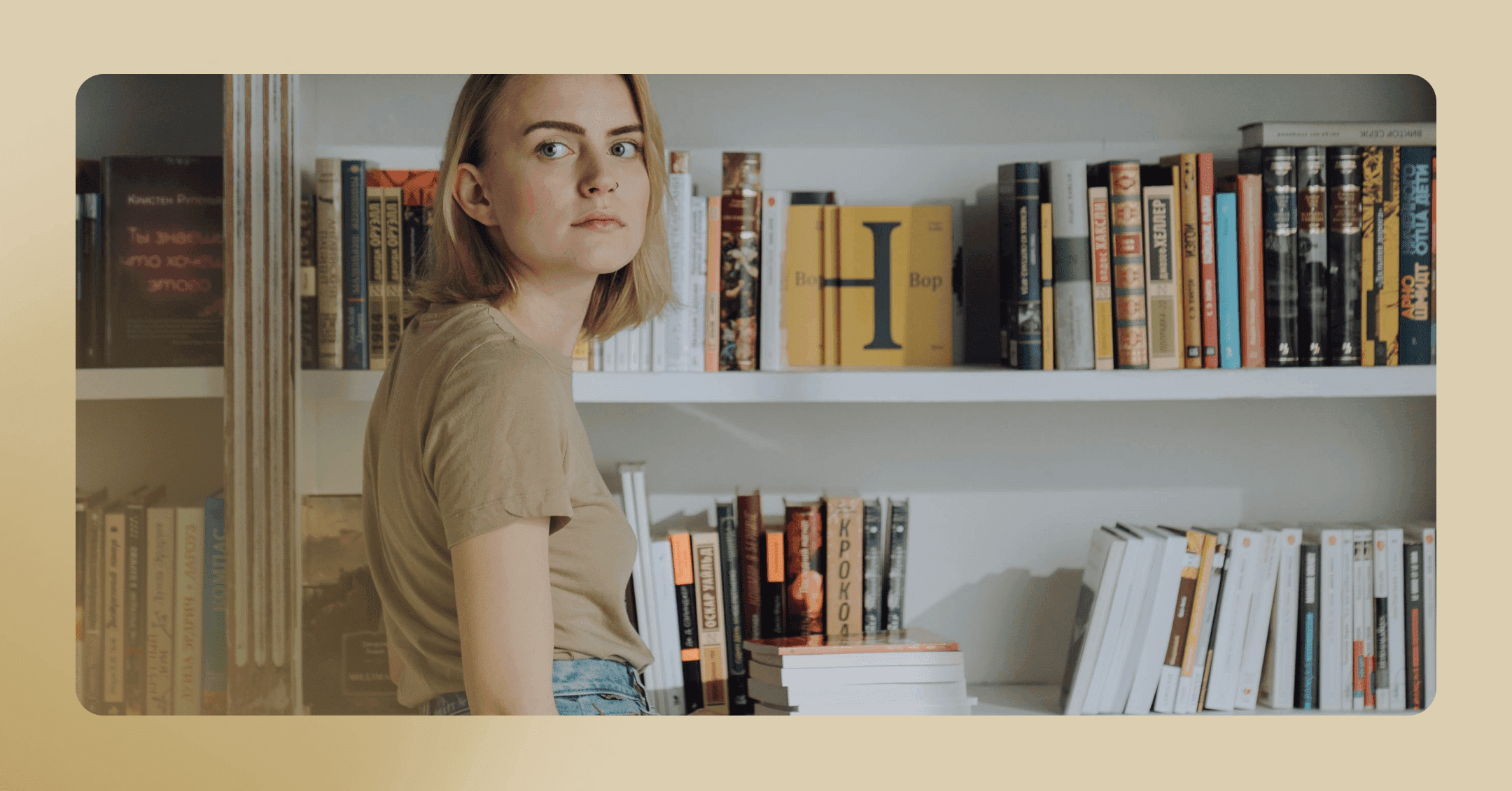 A woman with mid-length brown hair stand in front of a bookcase while looking of to the side. She has a serious look on her face and is wearing a tan short sleeve shirt.