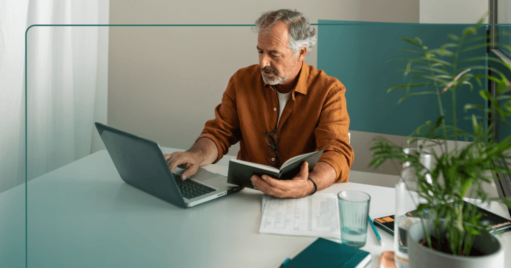 Older man sits at a table with papers, a near empty glass, and books. He is scrolling on an open laptop with his right hand and holds an open book with his left hand. He is wearing an orange long sleeve shirt. The background is a beige wall.