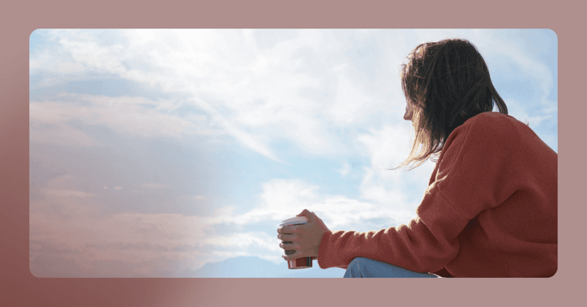 A woman in an orange sweatshirts holds a coffee to-go cup and looks of the frame to the left. A blue sky with clouds is visible in the background. The image is framed in a light red color.